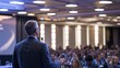 © VistaVisions - Speaker giving a talk at a corporate business conference. Audience in auditorium hall with presenter in front of presentation screen. Corporate executive giving speech during entrepreneur seminar
