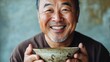 © Sinechana - 26.A portrait of an Asian man with a cheerful smile, holding an empty ceramic bowl with both hands, captured against a softly lit neutral background.
