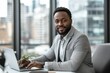 © SimpleDesignStudio - Confident Young Black Man in Professional Attire Working Remotely at Modern Office Desk with Laptop Amidst Cityscape Background