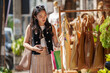 © crizzystudio - Stylish young woman holding her smartphone and shopping bags is choosing reusable handmade bags at an outdoor market stall promoting sustainable and eco-conscious lifestyle