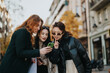 © qunica.com - Young women enjoying a fun moment outside in a city setting, sharing laughs as they look at a smart phone together, capturing a cheerful and connected atmosphere among friends.