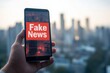 © alisluch - Hand holding a smartphone displaying a fake news warning against a blurred cityscape, symbolizing misinformation, digital media, and online deception