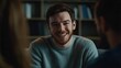 © Elena - Young man with beard sitting at table, smiling and laughing. He is in a friendly discussion with people around him. The setting appears to be indoors, possibly a café or office.