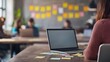 © somchairakin - A person sits at a desk with a laptop and sticky notes, while others collaborate in a modern workspace filled with colorful post-its.