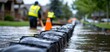 © apichat - The image shows workers in safety vests managing flooding, with sandbags and a flooded street in a residential area.