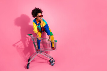 Wall Mural - Stylish woman in vibrant clothing posing with shopping cart against a pink background