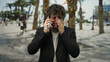 © Krakenimages.com - Young man crying while talking on cellphone outdoors on a city street, with blurred palm trees and pedestrians in the background, depicting emotional expression.