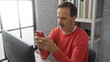 © Krakenimages.com - Mature man wearing red sweater uses smartphone indoors at office, looking thoughtful while sitting at desk surrounded by books and facing a large window in a well-lit interior space.