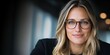 © imagineRbc - A confident woman with long, wavy hair and stylish glasses is smiling while seated in a contemporary office. The background has soft lighting and a blurred effect, creating a warm atmosphere