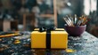 © JoxyAimages - A delightful yellow present tied with a black ribbon is placed on an artist's table full of vibrant colors and brushes, invoking feelings of creativity and inspiration.
