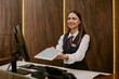 © AnnaStills - Medium close up of smiling young female receptionist in smart clothes standing behind reception desk with computer, telephone and payment terminal and holding white tablet