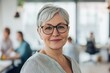 © Enigma - Portrait of a successful mature businesswoman with eyeglasses smiling confidently in a blurred modern office environment, colleagues working in the background