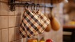 © Muekdum Studio - Warm Kitchen Scene: Close-up of oven mitts hanging on a rack with checkered pattern in a cozy kitchen setting, bathed in warm, natural light.