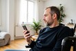 © Nina - Young Adult Caucasian Man in a Wheelchair Engaged with His Smartphone While Sitting in a Modern, Bright Living Room Filled with Greenery