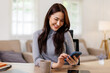 © David - Caucasian young asian woman sits at table uses mobile phone smiles at home office  Girl lady texting share messages content on smartphone social media applications online watching relax movie.
