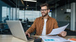 © dubogray1985 - The photo shows a smiling man with a beard and glasses sitting at a wooden desk in a modern office. He is wearing a brown jacket over a white shirt and is working on a laptop