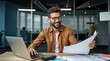 © dubogray1985 - The photo shows a smiling man with a beard and glasses sitting at a wooden desk in a modern office. He is wearing a brown jacket over a white shirt and is working on a laptop