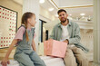 © DragonImages - Wide shot of father and daughter sitting on white bench while chatting about day spent in shopping mall