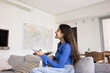 © fizkes - Young 20s Indian woman, owner or renter of modern smart home, resting alone seated on sofa holds remote control, switching on air conditioner, cooling or warming living room. Climate control, comfort
