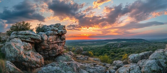 Poster - Stunning sunset panorama over rocky terrain with vibrant orange and blue hues illuminating the sky and lush green valleys in the background.