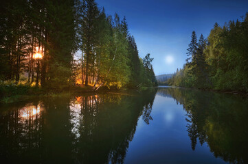 Wall Mural - A moonlit forest, with a campfire beside a calm river, and the sun setting behind the trees.