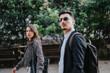 © qunica.com - A man and woman in business attire walk through a vibrant city park. The image captures a moment of casual conversation and modern urban style.