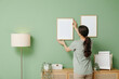 © Pixel-Shot - Young pretty woman hanging picture on green wall near wooden chest of drawers at home
