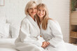© Pixel-Shot - Happy young woman and her mother in bathrobes sitting on bed at home