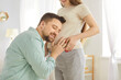 © Studio Romantic - Happy loving husband listening to baby in pregnant wife belly in the living room at home. Young couple and future parents expecting a baby. Parenthood, maternity and pregnancy concept.