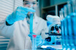 © Dino - Woman in a lab coat is holding a beaker of blue liquid. She is wearing gloves and a lab coat