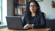 © BURINKUL - Latin woman business leader sitting at a wooden desk, laptop open, focused on strategy planning