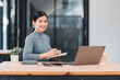 © PRIME STOCK LAB - Smiling Asian businesswoman working at desk, holding smartphone and pointing at laptop screen in bright office.