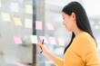© kenchiro168 - Asian businesswoman in yellow sweater smiles while organizing sticky notes on glass board, holding notebook.
