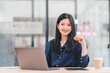 © kenchiro168 - Confident Asian businesswoman in striped shirt working at desk with laptop, tablet, and coffee cup in modern office.