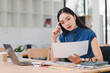 © kenchiro168 - Asian businesswoman in blue top sits at desk, looking stressed while using calculator, surrounded by office supplies.