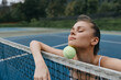 © SHOTPRIME STUDIO - Young athletic woman enjoying a moment of relaxation with a tennis ball on her shoulder, set on a serene blue tennis court surrounded by greenery