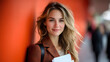 © Alena - A young blonde Caucasian businesswoman in a stylish orange blazer standing against a bold orange background, smiling confidently while holding documents