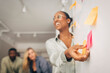 © Maskot - Smiling young businesswoman holding adhesive note on whiteboard during discussion in meeting room at office