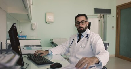 Serious young Hispanic doctor man in glasses and white coat working at computer in clinic office, typing medical reports on keyboard, consulting paper documents on workplace table