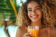 © imagemir - Happy young woman with curly brown hair enjoying a refreshing orange drink in a tropical setting.