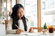 © bongkarn - An asian woman is smiling while writing in the notebook, sitting at the table beside window walls.
