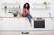 © sofiko14 - African woman sitting on kitchen counter operating robotic vacuum cleaner, looking happy and relaxed. Bright, clean kitchen environment with modern appliances showcases convenience.
