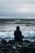 © BrandwayArt - Individual seated on rocks by the turbulent sea during cloudy weather