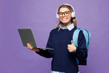 Young girl IT student wear blue casual clothes glasses headphones backpack bag hold use work on laptop pc computer show like isolated on plain purple background High school university college concept
