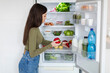 © Home-stock - Healthy eating concept. Rear view of young woman taking sweet pepper from refrigerator, searching vegetables for preparing meal