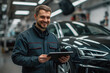© Jam - Mechanic in a car service center smiles while checking information on his tablet, surrounded by vehicles and tools in a busy environment