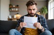 © MT - A thoughtful young Caucasian man with a beard, sitting on a sofa in a cozy living room, carefully reading a letter under soft ambient light, expressing concern or contemplation.