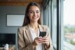 © MT - Smiling Young Woman in Professional Attire Holding a Smartphone in Modern Office Environment with Large Windows Overlooking a Scenic View