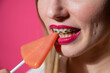 © Михаил Решетников - Portrait of a young woman with braces and bright makeup eating a lollipop on a pink background.