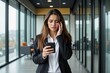 © MT - Young Latina Businesswoman in Black Suit Stressfully Reviewing Notifications on Smartphone Inside Modern Office Corridor with Colleagues in Background